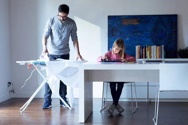 Jolie petite fille dessinant sur carnet tandis que son père repasse une chemise à la maison . — Photo