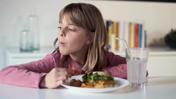 Vervelen klein meisje kijken opzij terwijl het eten van een volle hamburger met aardappelen thuis. — Stockvideo