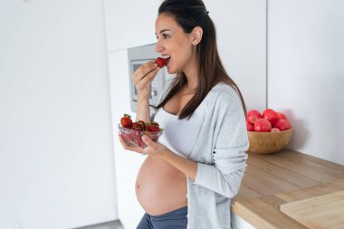 Güzel hamile genç kadın evde mutfakta kase çilek yemek.