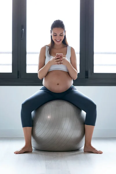 Mujer embarazada joven usando su teléfono móvil mientras hace ejercicio de relajación con pelota de fitness en casa . — Foto de Stock