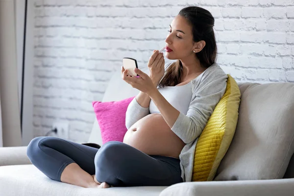 Beautiful pregnant young woman doing her makeup while sitting on sofa at home. — Stock Photo, Image