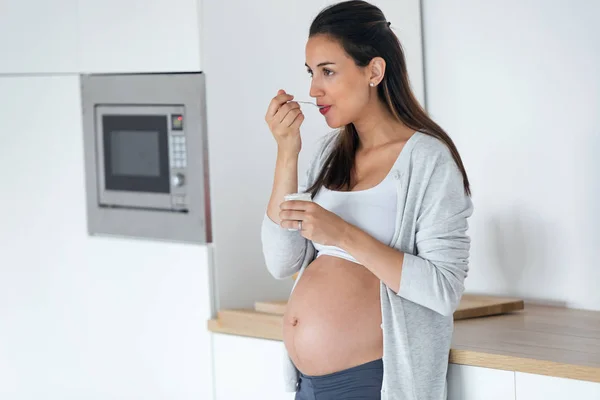 Beautiful pregnant young woman eating yogurt while standing in the kitchen at home. — Stock Photo, Image