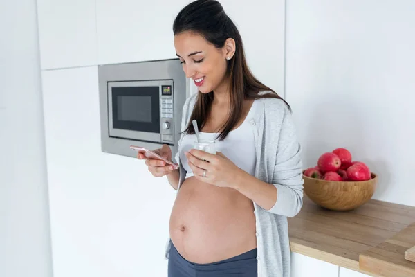Mujer joven bastante embarazada usando su teléfono móvil mientras come yogur en la cocina en casa . —  Fotos de Stock