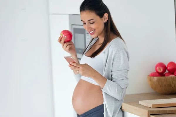 Mujer joven bastante embarazada usando su teléfono móvil mientras come una manzana en la cocina en casa . —  Fotos de Stock