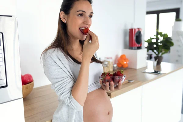Mooie zwangere jonge vrouw die aardbeien uit de kom eet in de keuken thuis. — Stockfoto