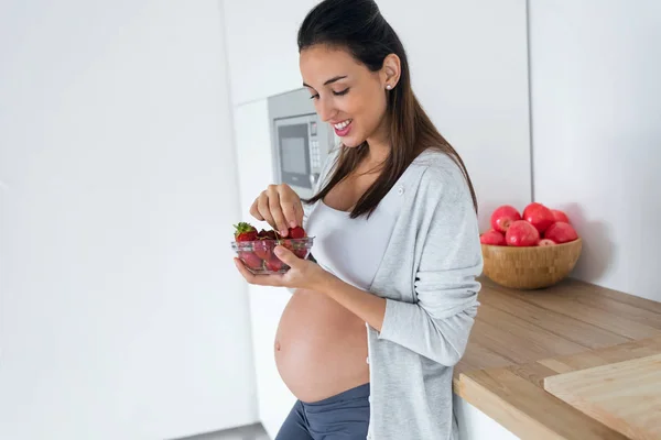 Gyönyörű terhes fiatal nő étkezési eper ki tálból a konyhában otthon. — Stock Fotó