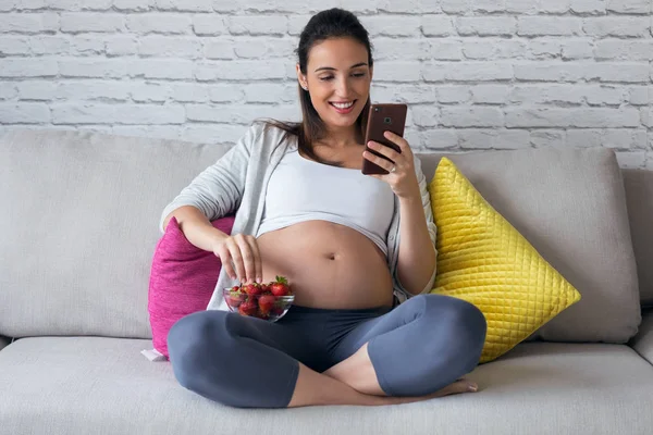Hermosa mujer embarazada usando su teléfono móvil mientras come fresas en el sofá en casa . —  Fotos de Stock