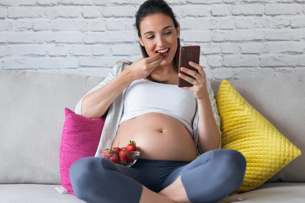 Hermosa mujer embarazada usando su teléfono móvil mientras come fresas en el sofá en casa . —  Fotos de Stock