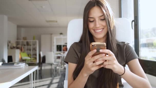 Video o usměvavé mladé obchodní ženě, která při sezení v kanceláři odesílá zprávy mobilním telefonem.