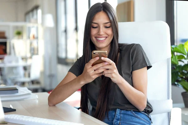 Leende ung affärskvinna skicka meddelanden med mobiltelefon när du sitter på kontoret. — Stockfoto