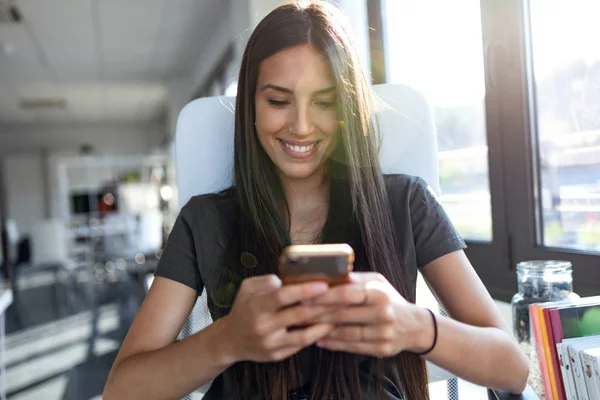 Leende ung affärskvinna skicka meddelanden med mobiltelefon när du sitter på kontoret. — Stockfoto