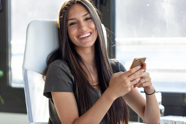 Lachende jonge zakelijke vrouw die naar de camera kijkt tijdens het gebruik van haar mobiele telefoon op kantoor. — Stockfoto