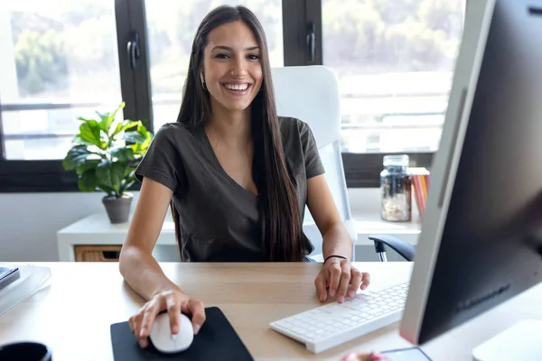 Sonriente joven mujer de negocios mirando a la cámara mientras trabaja con la computadora en la oficina . —  Fotos de Stock