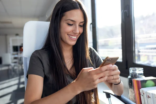 Lächelnde junge Geschäftsfrau, die im Büro Nachrichten mit dem Handy verschickt. — Stockfoto