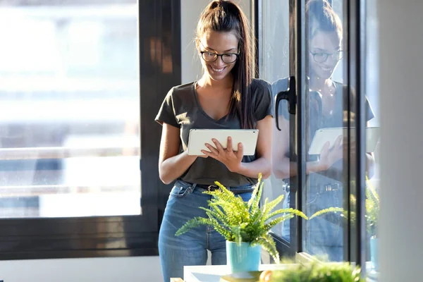 Uśmiechnięta młoda kobieta biznesu za pomocą swojego cyfrowego tabletu stojąc obok okna w biurze. — Zdjęcie stockowe