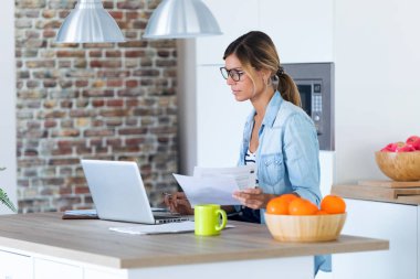 Evde mutfakta dizüstü bilgisayar ve belgelerle çalışan güzel genç kadın