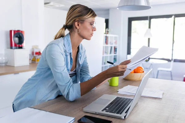Pretty young woman working with laptop and documents in the kitchen at home. — Stock Photo, Image