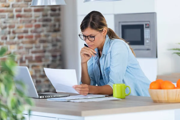 Jolie jeune femme travaillant avec un ordinateur portable et des documents dans la cuisine à la maison . — Photo