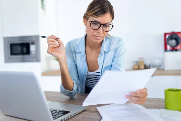 Jolie jeune femme travaillant avec un ordinateur portable et des documents dans la cuisine à la maison . — Photo