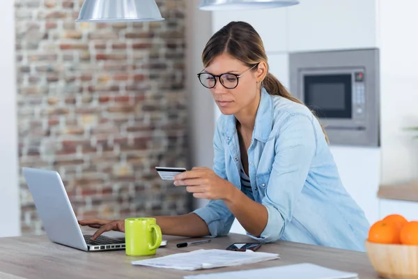 Ładna młoda kobieta za pomocą laptopa na zakupy online i płacąc kartą kredytową, siedząc w kuchni w domu. — Zdjęcie stockowe