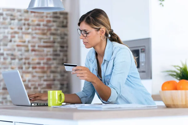 Mulher muito jovem usando seu laptop para fazer compras on-line e pagar com cartão de crédito enquanto está sentado na cozinha em casa . — Fotografia de Stock