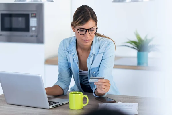 Pretty young woman using her laptop for shopping online and paying with credit card while sitting in the kitchen at home. — Stock Photo, Image