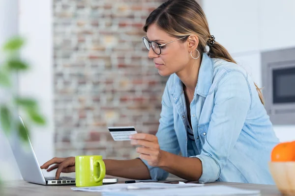 Mulher muito jovem usando seu laptop para fazer compras on-line e pagar com cartão de crédito enquanto está sentado na cozinha em casa . — Fotografia de Stock