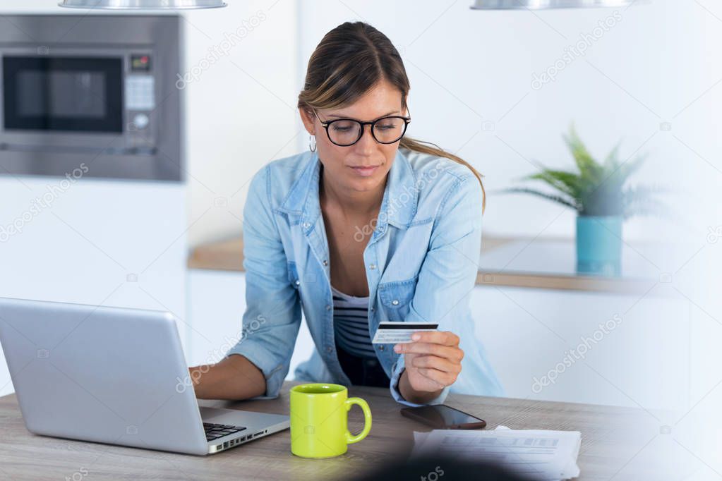 Pretty young woman using her laptop for shopping online and paying with credit card while sitting in the kitchen at home.