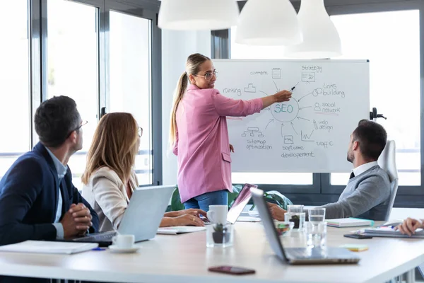 Elegantă tânără femeie de afaceri arătând spre tabla albă și explicând un proiect colegilor ei la locul de coworking . — Fotografie, imagine de stoc