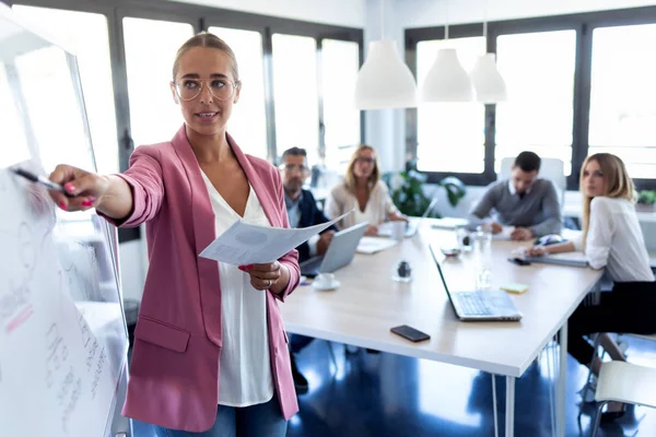 Elegancka młoda businesswoman wskazując na białym tablicy i wyjaśnić projekt do swoich kolegów na Coworking miejsce. — Zdjęcie stockowe
