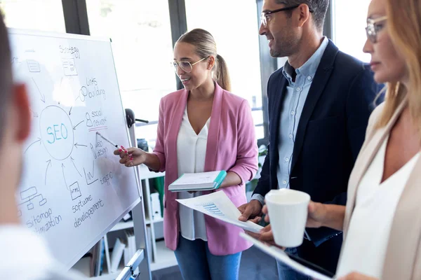 Elegante joven empresaria apuntando a pizarra blanca y explicar un proyecto a sus colegas en el lugar de coworking . — Foto de Stock