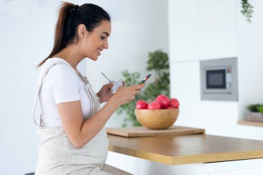 Evde mutfakta dondurma yerken onun cep telefonu kullanarak güzel genç hamile kadın.