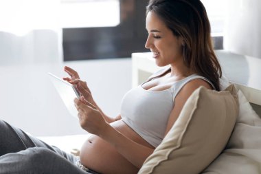 Evde yatakta otururken onu dijital tablet kullanarak güzel genç hamile kadın.