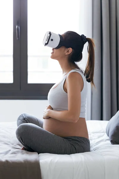 Gyönyörű fiatal terhes nő használ vele virtuális valóság szemüveg ülve az ágyon otthon. — Stock Fotó
