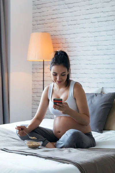 Hermosa joven embarazada usando su teléfono móvil mientras come cereales y se sienta en la cama en casa . — Foto de Stock