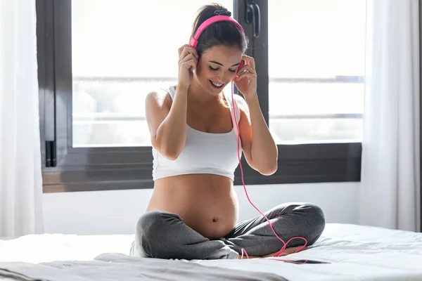 Piękna młoda kobieta w ciąży słuchając muzyki ze słuchawkami siedząc na łóżku w domu. — Zdjęcie stockowe