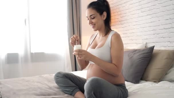 自宅のベッドに座りながらヨーグルトを食べる美しい若い妊婦のビデオ — ストック動画