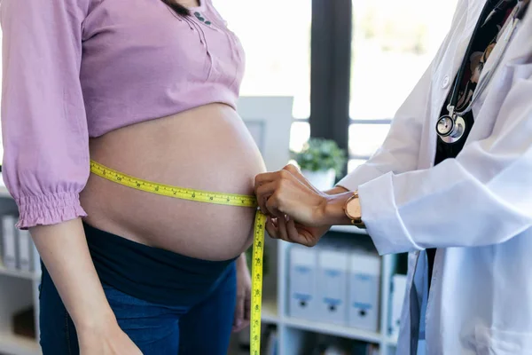 Pregnant woman exposing her belly while doctor using a measuring tape to follow the growth of the baby at a hospital.