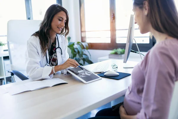 Mooie jonge vrouw gynaecoloog met behulp van digitale tablet voor het tonen van de echografie naar haar zwangere patiënt in de kliniek. — Stockfoto