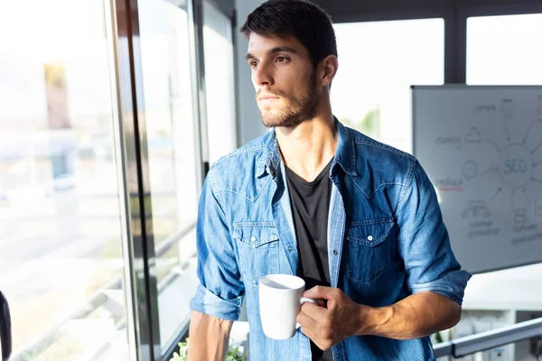Jovem empresário pensativo olhando pela janela enquanto bebe café e faz uma pausa no escritório . — Fotografia de Stock