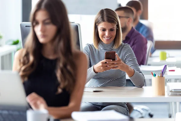 Sorrindo jovem empresária usando seu smartphone enquanto trabalhava com computador no escritório de inicialização moderna . — Fotografia de Stock