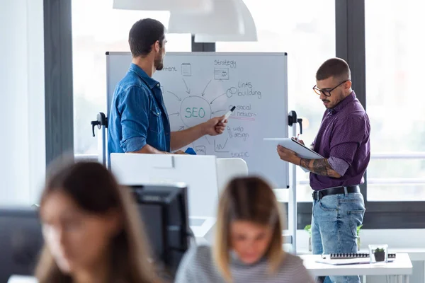 Jeune homme d'affaires pointant sur tableau blanc tout en discutant avec son collègue de leur nouveau projet dans le bureau de démarrage moderne . — Photo