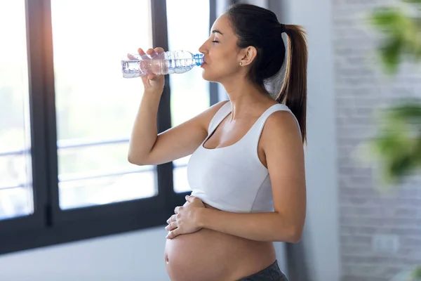 Pretty young pregnant woman drinking water while standing at home. — Stock Photo, Image