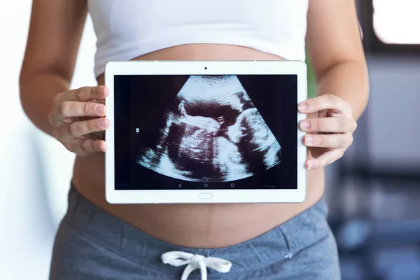 自宅に立っている間、胎児超音波写真でデジタルタブレットを保持し、示す妊婦. — ストック写真