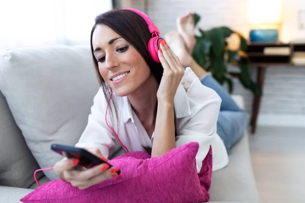 Güzel genç kadın kulaklık ile müzik dinlerken ve evde bir kanepede istirahat ederken cep telefonu kullanarak. — Stok fotoğraf
