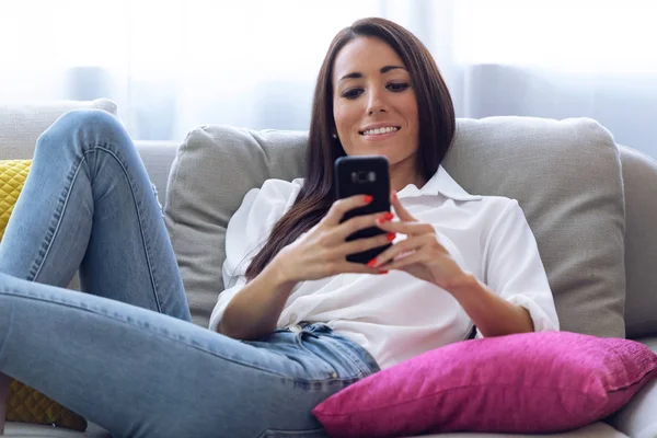 Mooie jonge vrouw met behulp van haar mobiele telefoon zittend op de Bank thuis. — Stockfoto