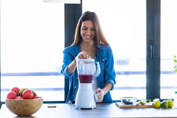 Bella giovane donna che mescola frutta nel frullatore per la colazione fresca in cucina a casa . — Foto Stock