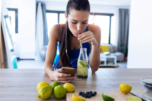 Hermosa joven deportista bebiendo jugo de desintoxicación mientras escucha música con teléfono inteligente en la cocina . — Foto de Stock