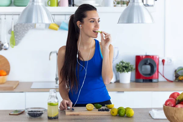 Mujer joven deportiva comiendo rodaja de lima mientras prepara bebidas desintoxicantes en la cocina en casa . — Foto de Stock