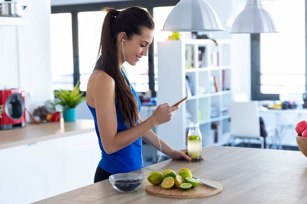 Sporty young woman listening to music with mobile phone after training in the kitchen at home. — Stock Photo, Image